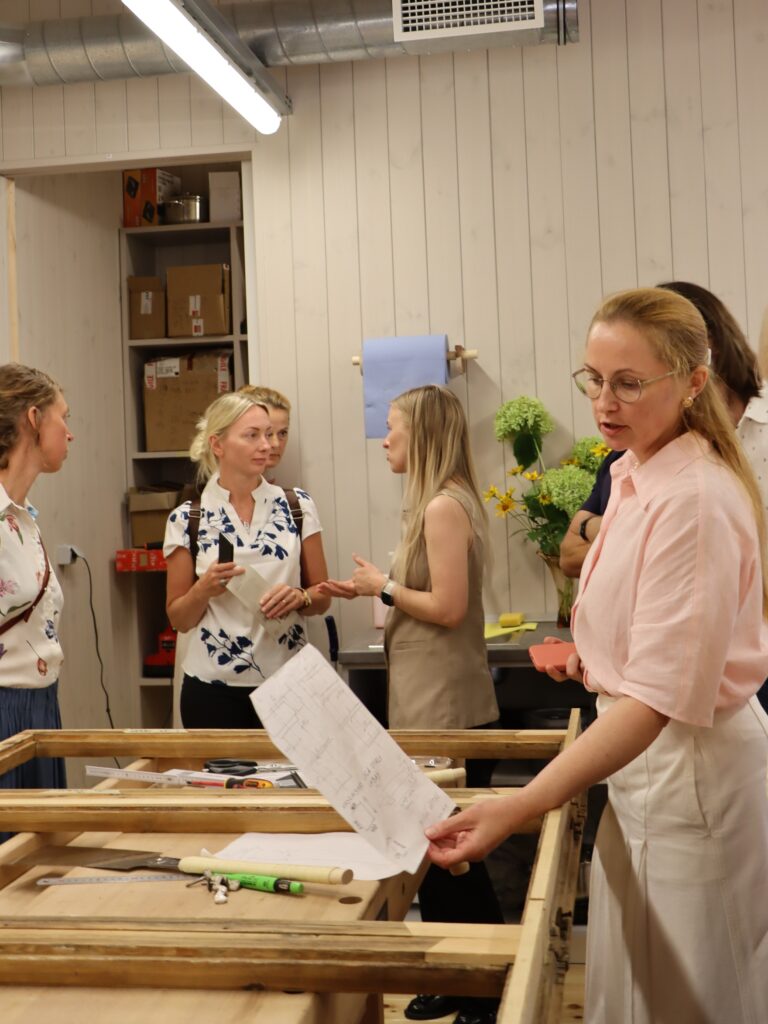 Grupa cilvēku darbnīcas vai mācību telpā pulcējas ap koka galdu ar instrumentiem un papīriem; viena persona tur lapu un, iespējams, kaut ko skaidro. Fonā redzamas kastes, plaukti un ziedi uz letes